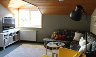 A cozy living room with a colorful sofa and a yellow armchair. The room has large windows that let in plenty of light and a modern TV corner.