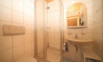A small bathroom with a shower, a sink, and a toilet. The walls are tiled and there is a window with natural light.