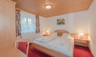 A cozy bedroom with two beds and wooden details. The walls are light, and there are windows with curtains and a small picture on the wall.