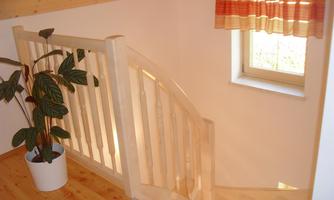 A bright staircase with a wooden railing leads downwards. Next to it, there is a plant in a white pot in front of a window with a colorful curtain.