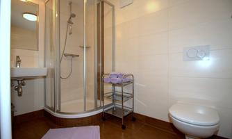 A modern bathroom with a shower and a toilet. There is a sink and a small rolling cart with towels.