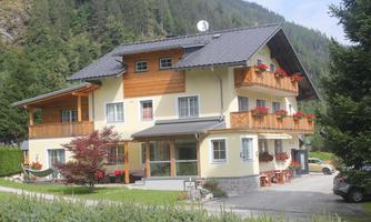 A charming building with wooden cladding and balconies, surrounded by trees and mountains. There is a well-maintained garden and a parking lot in front of the house.