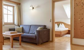 A cozy living room with a sofa and a table. In the background, there is a bedroom with a double bed and wood paneling visible.