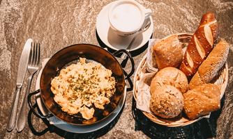 A plate with scrambled eggs in a pan, garnished with herbs. Accompanied by a selection of rolls and a cup of coffee.