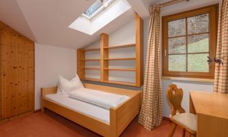 A simple room with a single bed and a desk. Large windows and shelves create a bright and friendly atmosphere.