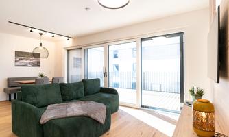 A modern living room with a green sofa and large windows. The room features light wooden flooring and a relaxing atmosphere.