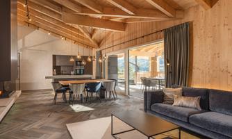 A modern living room with a wooden ceiling and large windows. The space is bright and inviting, furnished with a sofa and a dining table.