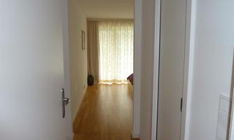 A bright hallway with wooden flooring leads to a room with large windows and curtains. There is a calm and inviting atmosphere.