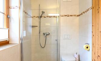 A modern bathroom with a glass shower cubicle and a wall-mounted toilet. The walls are brightly tiled and a window provides daylight.