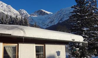 A snowy mountain landscape with a house in the foreground. The clear blue sky and the majestic mountains create a calm atmosphere.