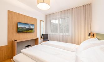A cozy hotel room with two single beds and a television mounted on the wall. Large windows provide plenty of daylight and a pleasant atmosphere.