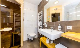 A modern bathroom with a shower and toilet area. It features a white sink and a wooden shelf.