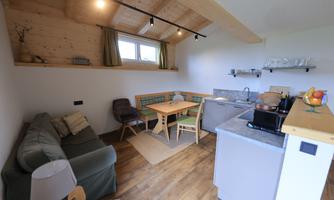 A cozy living kitchen with a wooden ceiling, a dining table, and a small kitchenette. The room is bright and furnished in a modern style.