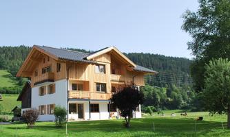 A beautiful house in Alpine style with wood paneling. It is situated in a green landscape with mountains in the background.
