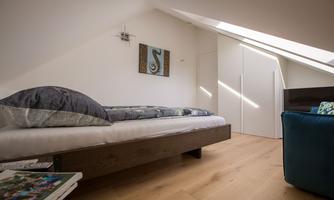 A simple bedroom with a bed and wooden floor. Light falls through a skylight, creating a bright atmosphere.