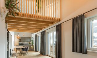 A modern, spacious living room with wooden flooring and large windows. The wooden ceiling construction and elegant lamps create an inviting atmosphere.