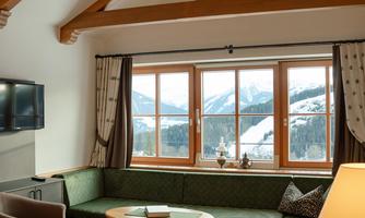 A cozy living space with a green couch and large windows that offer a view of the mountains. The room has a modern design with wooden beams and warm lighting.