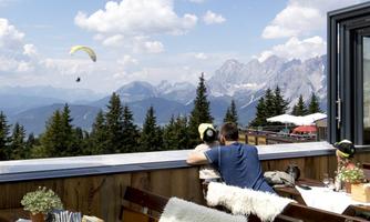A restaurant with a beautiful view of the mountains and forests. In the background, you can see a paraglider in the sky.