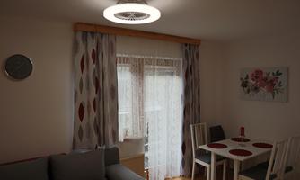 A cozy living room with a sofa and a small dining table. The room is brightly lit and has decorative curtains as well as a wall painting.