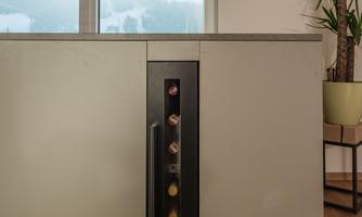 A modern kitchen unit with an integrated wine cooler. In the background, there are windows with a mountain view and a plant visible.