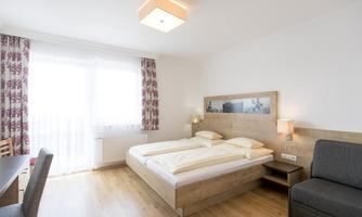 A modern hotel room with a double bed, a cozy armchair, and a desk. Large windows provide plenty of daylight and a pleasant atmosphere.