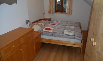 A cozy bedroom with a double bed and wooden furniture. The room has a window with curtains and a warm wooden floor.