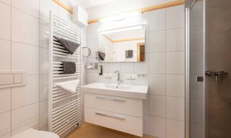 A modern bathroom with a white washbasin and a mirror. Towels hang on a towel warmer, and a shower is visible.