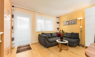 An inviting living room with a gray sofa, a small table, and plants. Large windows let in plenty of light, and the walls are painted in warm shades of yellow.