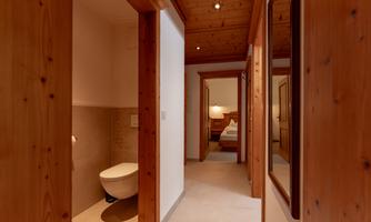 A bright hallway with a wooden ceiling and walls. On the left side, there is a bathroom with a toilet, while in the background, a bedroom is visible.