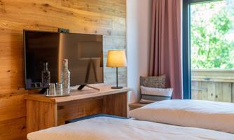 A modern bedroom with two beds, a television, and a wooden wardrobe. A lamp and a window overlooking the outdoors complete the cozy atmosphere.