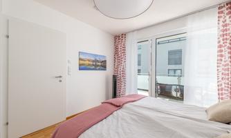 A modern bedroom with a double bed and a soft, pink bedspread.  
Large windows let in plenty of daylight and offer a view of the balcony.