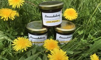 Three jars of dandelion honey are standing on a green meadow, surrounded by bright yellow dandelion flowers. The jars have simple labels that read "Dandelion Honey."