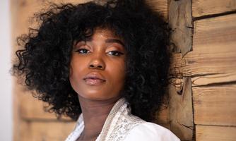 A young woman with full, curly hair is looking directly at the camera. In the background, a rustic wooden wall can be seen.