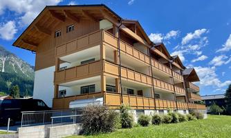 A modern wooden building with several balconies under a blue sky. Surrounded by green fields and mountains.
