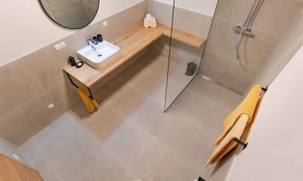 A modern bathroom with a glass shower cubicle and a wooden bench. There is a round mirror and toiletries on the sink.