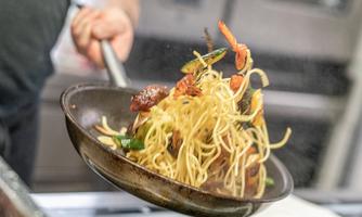 A chef tosses a pan with stir-fried noodles and colorful vegetables. The dishes dance in the air, showcasing a vibrant cooking scene.