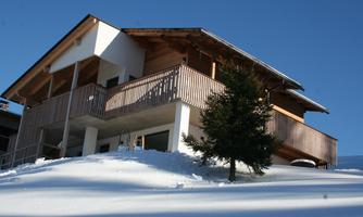 A modern house with wooden cladding stands on a snow-covered hill. The clear blue sky provides a fresh winter atmosphere.