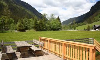 A beautiful wooden terrace with seating offers a view of green meadows and surrounding mountains. The sky is partly cloudy and it is a peaceful landscape.