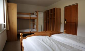 A simple room with two beds and a bunk bed in the background. There is a wooden cupboard and a door that leads outside.