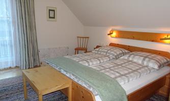 A cozy bedroom with a double bed and wooden details. A table and a chair complete the furnishings, while bright curtains let light in.