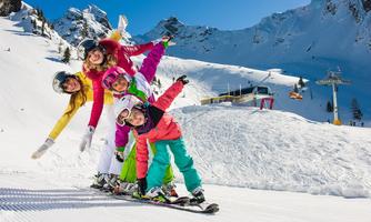A group of cheerful skiers stands on their skis in a snow-covered ski area. In the background, mountains and a ski lift station can be seen.
