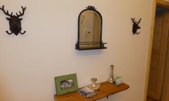 A simple hallway with a wooden wall shelf, a mirror, and a few decorative items. Two stylized antler heads are hanging on the wall.