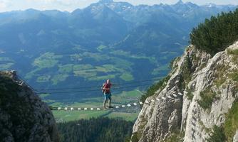 A hiker stands on a hanging bridge between two rocks. In the background, impressive mountains and a green landscape can be seen.