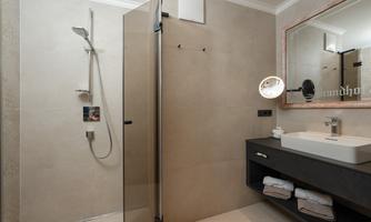 A modern bathroom with a glass shower and a sink on a black vanity. In the background, a illuminated mirror reflects, and fresh towels are visible.