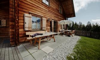 A rustic wooden house with a cozy terrace. On the terrace, there is a table and chairs, surrounded by a beautiful green meadow.