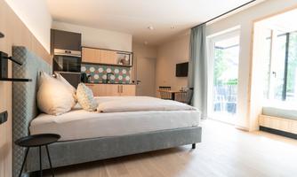 A modern room with a large bed and a small kitchenette. The large windows provide plenty of light and a view outside.