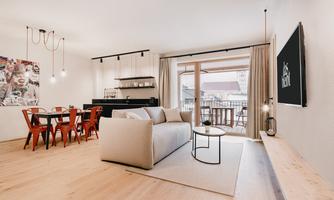 A modern living room with a comfortable couch and a wooden table. The room has large windows leading to a balcony, as well as an adjacent dining area.