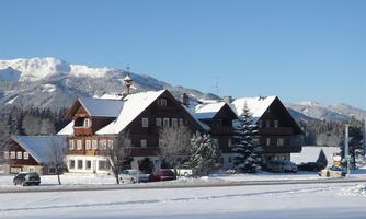 A snowy landscape with a traditional wooden building and mountains in the background. The sky is clear and blue.