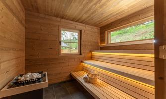 A modern sauna with wood paneling and soft lighting. There are wooden benches and a small window with a view of the greenery.