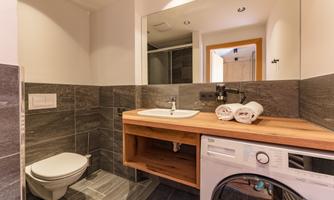 A modern bathroom with dark tiles and a wooden shelf. It includes a sink, a toilet, and a washing machine.